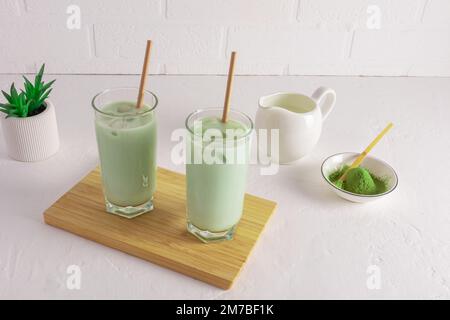 due bicchieri di tè matcha e latte appena fatti su un asse di legno su un tavolo bianco contro una parete di mattoni bianchi. un'alternativa al caffè del mattino Foto Stock