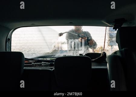 Autolavaggio uomo con pistola ad acqua in autolavaggio self-service. Sapone, cera e gocce d'acqua che coprono il cristallo del finestrino del veicolo. Visto dall'interno dell'autom Foto Stock