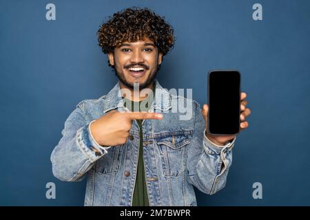 Mock-up, copia spazio. Felice eccitato indiano o arabo ragazzo, in abbigliamento casual, tiene smartphone con schermo bianco vuoto per la presentazione e punta dito Foto Stock