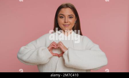 Donna innamorata. Donna sorridente 20s in maglione bianco fa il gesto del cuore dimostra segno d'amore esprime buoni sentimenti e simpatia. Giovane bella ragazza adulta isolata da sola su sfondo rosa studio Foto Stock