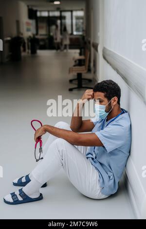 Giovane medico in difficoltà seduto al pavimento del corridoio dell'ospedale. Foto Stock