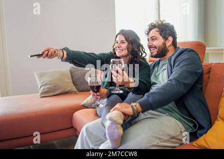 Coppia amorevole bere vino e guardare la tv seduto sul divano e tenendo un telecomando - sposi novelli divertirsi nel fine settimana a casa guardando taltezza Foto Stock