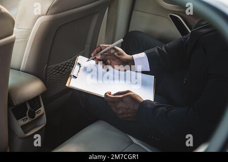 Concetto aziendale di un bell'uomo dalla pelle scura in tuta e occhiali nella seduta posteriore di un'auto di lusso che lavora e prende appunti. Firmare i documenti. chiudi Foto Stock