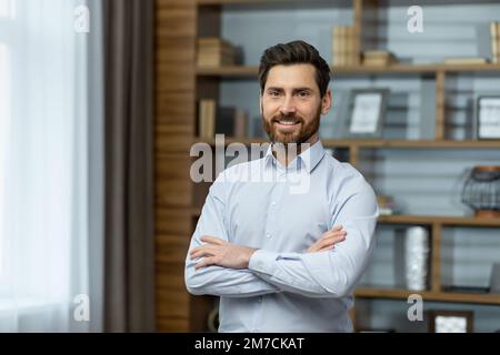 Ritratto di un uomo d'affari di successo in ufficio, uomo in camicia sorridente e guardando la macchina fotografica, boss maturo con barba con mani scoscese in piedi sul posto di lavoro all'interno dell'edificio. Foto Stock