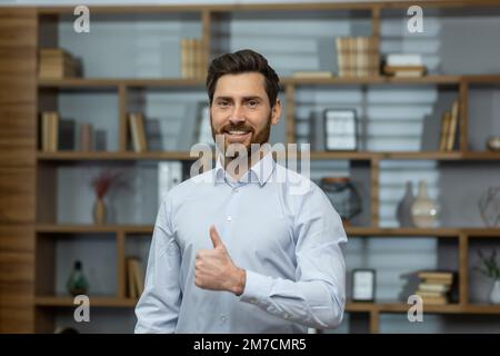 Uomo d'affari riuscito sorridente e guardando la macchina fotografica, l'uomo mostra i pollici in su affermativamente e gioiosamente concorda con il successo, il capo in camicia all'interno dell'ufficio al lavoro. Foto Stock