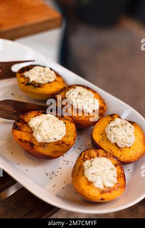 Gustose pesche tagliate alla griglia, servite con panna montata al formaggio, primo piano. Cibo delizioso, dessert. Festa all'aperto Foto Stock