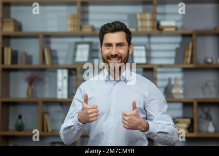 Uomo d'affari riuscito sorridente e guardando la macchina fotografica, l'uomo mostra i pollici in su affermativamente e gioiosamente concorda con il successo, il capo in camicia all'interno dell'ufficio al lavoro. Foto Stock