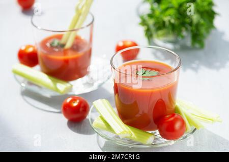 Due bicchieri con succo di pomodoro fresco, sedano, prezzemolo e pomodori maturi su sfondo di legno chiaro Foto Stock
