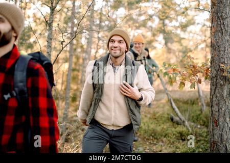 Uomo sorridente che indossa abiti caldi mentre fai escursioni con gli amici nella foresta Foto Stock