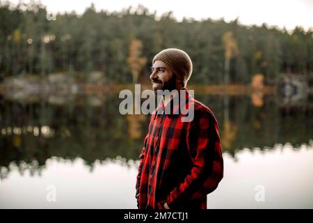 Uomo sorridente che indossa una maglietta a quadri mentre si trova vicino al lago Foto Stock