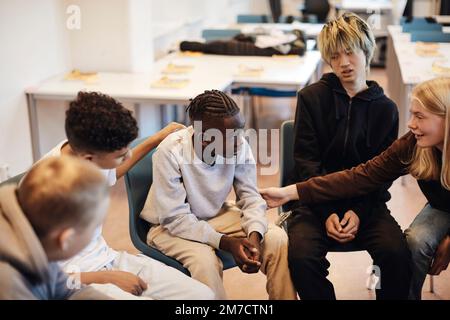 Ragazza adolescente bionda che consolava il ragazzo seduto tra amici maschi in classe durante la terapia di gruppo Foto Stock