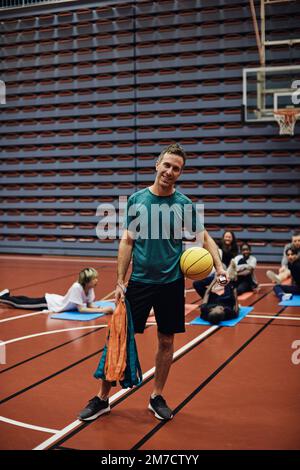 Ritratto completo di un allenatore maschile sorridente in piedi con basket contro gli studenti al campo sportivo Foto Stock