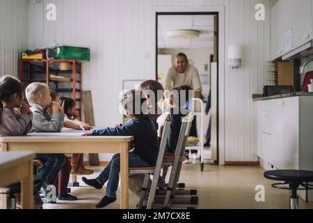 Studenti maschili e femminili multirazziali seduti al tavolo da pranzo durante la colazione nell'asilo Foto Stock