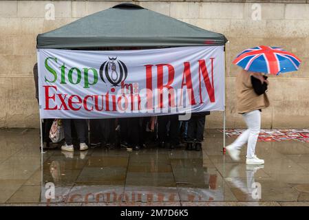 Stop esecuzione in Iran banner di protesta, sul gazebo in Trafalgar Square, Londra. Giorno piovoso, con i manifestanti al riparo. Persona con ombrello Union Flag Foto Stock