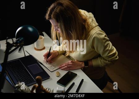 Donna messa a fuoco in abiti casual guardando lo schermo del notebook e disegno illustrazione in notebook. La freelance femminile lavora in un ambiente di lavoro domestico Foto Stock