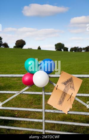Ballons legato con un cartello di cartone stabilito nel paese, il segno punta con una freccia e i ballon significa che un partito è in corso e indica la direzione in cui si sta tenendo. Foto Stock