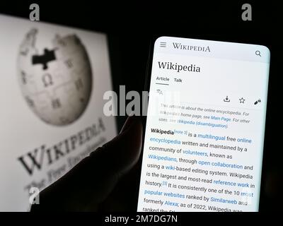 Persona che tiene il cellulare con la pagina web dell'enciclopedia online Wikipedia sullo schermo di fronte al logo. Messa a fuoco al centro del display del telefono. Foto Stock
