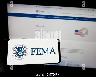 Persona che tiene lo smartphone con il sigillo della Federal Emergency Management Agency (FEMA) sullo schermo di fronte al sito Web. Messa a fuoco sul display del telefono. Foto Stock