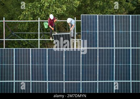 Lavoratori che detengono ed installano pannelli solari. Gli uomini indossano caschi e uniformi. Lavoro di squadra. Array come sistema di pannelli fotovoltaici. Array di un sistema a pannelli solari che fornisce energia solare. Foto Stock