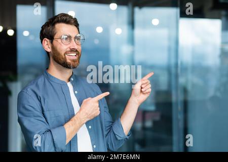 Uomo d'affari sorridente all'interno dell'ufficio che punta le dita alla direzione, capo felice in camicia e occhiali. Foto Stock