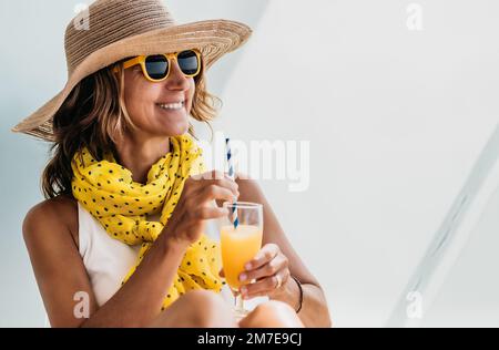 primo piano di una donna di mezza età con un cappello e bicchieri gialli che tiene in mano un succo d'arancia. concetto di vacanza e relax. Foto Stock