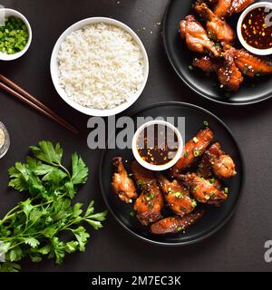 Ali di pollo alla soia e miele appiccicose e ciotola di riso su fondo di pietra scura. Vista dall'alto, disposizione piatta Foto Stock