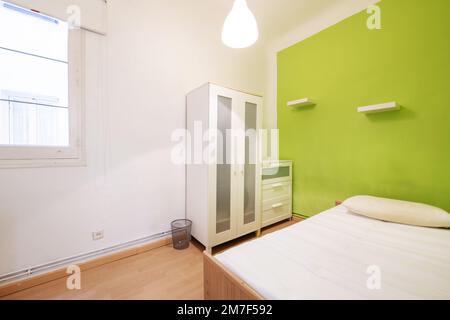 Camera da letto con un ampio letto, una parete dipinta di verde pistacchio, un armadio bianco e una cassettiera e una finestra in legno con una tenda Foto Stock