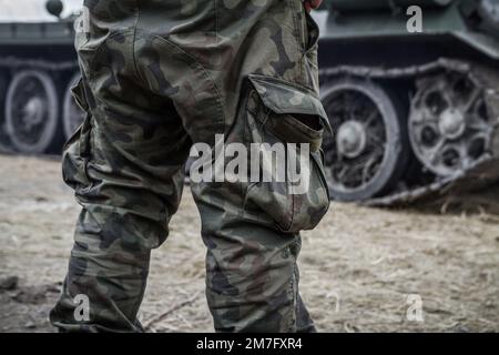 Gambe soldato su un campo di battaglia, indossando pantaloni mimicanti mimicanti in legno, pantaloni mimetizzazione. Carro armato militare cingolato ruote in background. Foto Stock