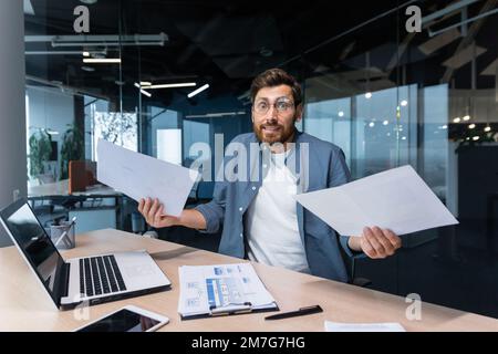 Insoddisfatto e arrabbiato uomo d'affari all'interno dell'ufficio che mostra rapporti e fatture documenti alla macchina fotografica, capo in camicia che guarda la macchina fotografica che lavora con il lavoro di carta del laptop. Foto Stock