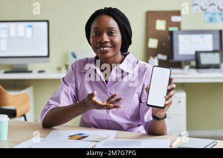 Ritratto di donna nera sorridente che presenta smartphone con schermo bianco mockup in ufficio designer e sorridente alla fotocamera, copia spazio Foto Stock