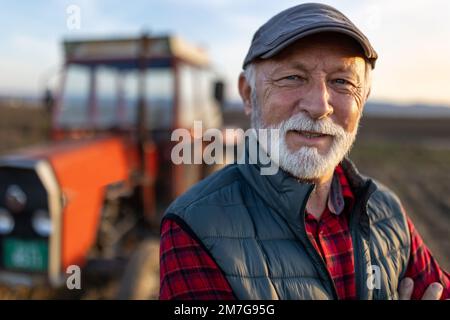Ritratto di contadino anziano con i capelli bianchi e la barba in piedi davanti al trattore sul campo a fine autunno Foto Stock