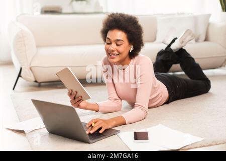 Felice giovane donna mista in cuffie wireless con computer e blocco note, si trova sul pavimento, godersi la videochiamata Foto Stock