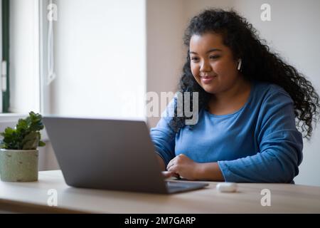 Giovane donna ispanica più taglia che usa laptop e auricolari in interni Foto Stock