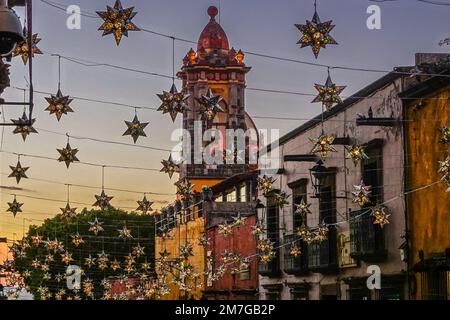 Colorate decorazioni a stella di stagno si stagliano in edifici storici con il campanile della Chiesa dell'Immacolata Concezione su Canal Street al crepuscolo nel quartiere storico, 4 gennaio 2023 a San Miguel de Allende, GTO, Messico. Foto Stock