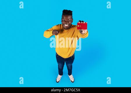 Cheerful Black Guy tenendo in mano la carta di credito e indicando la fotocamera Foto Stock