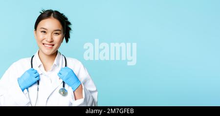 Medico donna asiatica, infermiere sorridente fiducioso, stetoscopio di regolazione sul collo, in piedi in uniforme medica su sfondo blu Foto Stock
