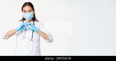 Cura medico asiatico, medico donna in maschera medica e guanti di gomma mostra la sua cura per i pazienti con segno di cuore, sfondo bianco Foto Stock