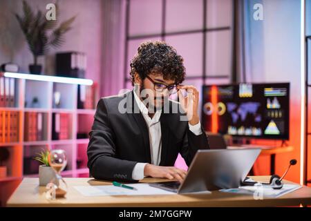 Uomo d'affari con esperienza, economista, borsone in tuta con occhiali seduti al tavolo con un notebook la sera in ufficio. Concetto di scadenza del progetto. Foto Stock