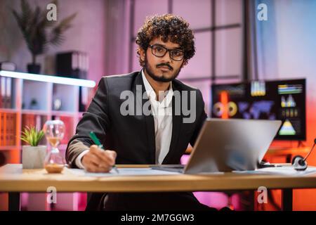 Ritratto di attraente sicuro barbuto specialista IT, Office manager, indossare tuta utilizzando il laptop fare report finanziario mentre si scrive su carta, guardando la fotocamera al lavoro di notte. Foto Stock