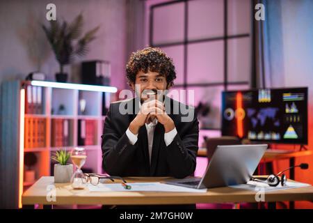 Uomo sorridente in abito formale seduto alla scrivania con un moderno computer portatile e guardando la fotocamera la sera in ufficio. Concetto di persone, scadenze e lavoro in ufficio. Foto Stock