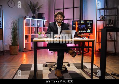 Ritratto completo di un uomo sorridente barbuto in tuta formale seduto alla scrivania con un moderno computer portatile e guardando la fotocamera la sera in ufficio. Concetto di persone, scadenze e lavoro in ufficio. Foto Stock