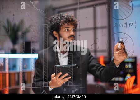 Primo piano attraverso la vista di vetro di focalizzato giovane uomo d'affari barbuto disegno grafico a torta su tavola di vetro, mentre lavorando in ufficio moderno di notte. Foto Stock