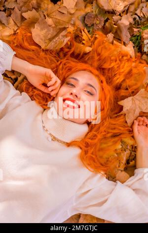 Donna rossa sdraiata sulle foglie nel parco cittadino sorridente, stile di vita autunnale Foto Stock
