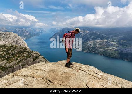 Escursionista in piedi con attenzione allo sperone roccioso, vista dallo sperone roccioso Preikestolen al Lysefjord e le montagne, tempo soleggiato, Ryfylke, Rogaland Foto Stock