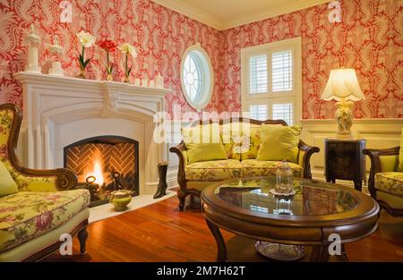 Caminetto a gas propano illuminato, poltrone e divano rivestiti di fiori gialli in salotto all'interno di un'elegante casa in stile cottage. Foto Stock