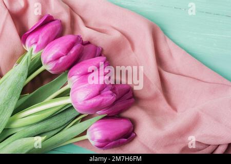Mazzo di tulipani viola su tessuto rosa su tavolo di legno verde. Concetto di vacanza per le donne. Vista dall'alto, disposizione piatta. Foto Stock