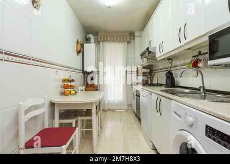 Cucina con mobili bianchi ed elettrodomestici di tutti i tipi e colori, piani di lavoro color crema e piastrelle abbinate, tavolo da cucina in legno bianco con cha Foto Stock