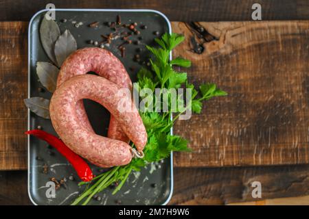 Anelli di salsiccia close-up.Raw anelli di salsiccia fatti in casa, prezzemolo fresco, peperoncino rosso in un piatto nero su un tavolo di legno scuro. appetitosi prodotti a base di carne Foto Stock