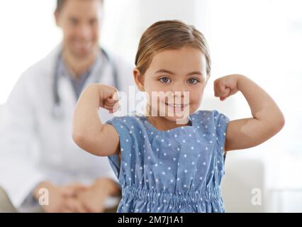 Ha mangiato tutte le sue verdure. Una bambina che flette i suoi muscoli con il medico sullo sfondo. Foto Stock