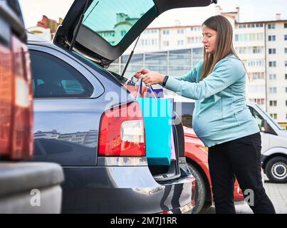 Vista laterale della donna caucasica con pancia puting borse shopping in bagagliaio auto godendo maternità e paternità in modalità attesa. Donna incinta che si prepara alla nascita del bambino. Foto Stock